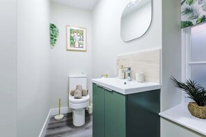 A bathroom with a toilet, sink, and mirror. There is a framed picture on the wall and a plant on the windowsill. - NEW! Modern 1-Bed Flat, Great Location, Sleeps 4! (Chester)