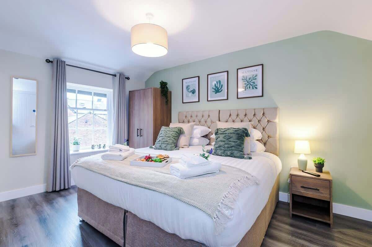 A well-lit bedroom with a large bed, a bedside table with a lamp, and framed artwork on the wall.
