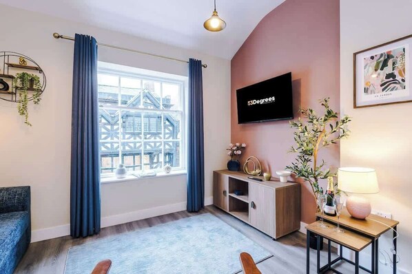 A modern living room with a large window, a flat-screen TV, a wooden cabinet, a small table with a lamp, and a blue sofa. - NEW! Modern 1-Bed Flat, Great Location, Sleeps 4! (Chester)