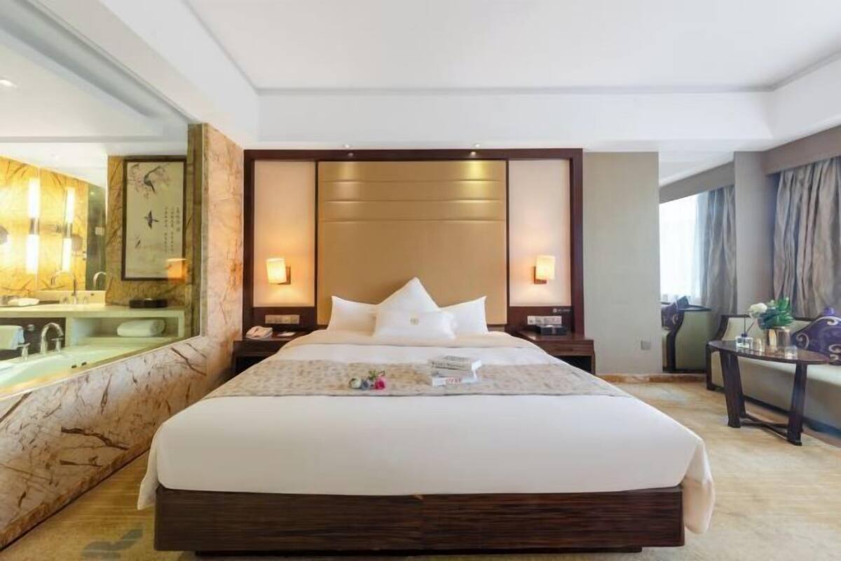 A hotel room with a large bed, a sitting area with a marble wall, a bathroom with a sink and mirror, and a dining area with a table and chairs.