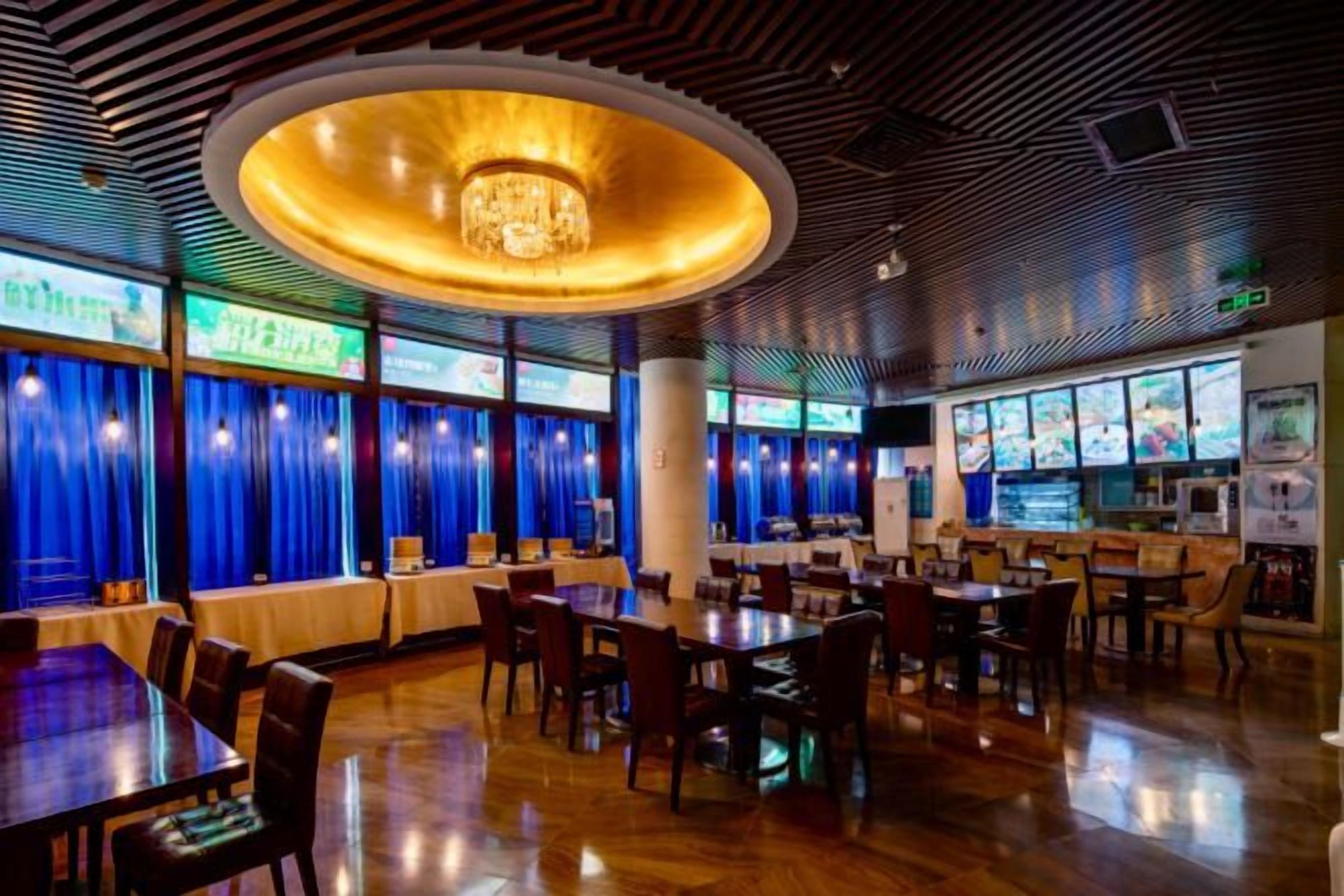 A restaurant interior with round tables, chairs, a central chandelier, and blue curtains.