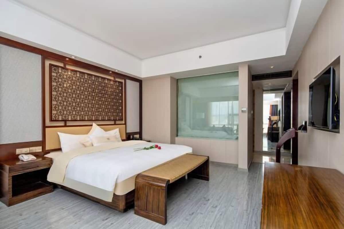 A modern hotel room with a large bed, a wooden bench, a nightstand, a TV mounted on the wall, and a large window.