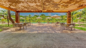 Un espace extérieur aménagé en terrasse, avec des tables et des chaises, une pergola au-dessus et une vue sur une piscine et de la verdure.