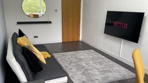 Una sala de estar moderna con un sofá gris, un sillón amarillo, un tapete gris y un televisor montado en la pared mostrando el logo de Netflix.