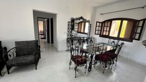 Un coin repas avec une table et des chaises en verre, un miroir décoratif et une vue sur un jardin par une fenêtre ouverte.
