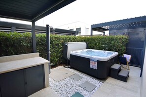 Uma banheira de hidromassagem sob um alpendre coberto, cercada por um sebedo e um deck revestido de azulejos.