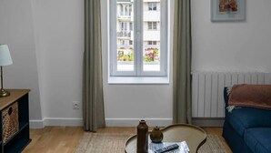 Un moderno salón con tragaluz, un ventanal con cortinas, un sofá azul, una pequeña mesa redonda con un jarrón y un control remoto, y piso de madera.