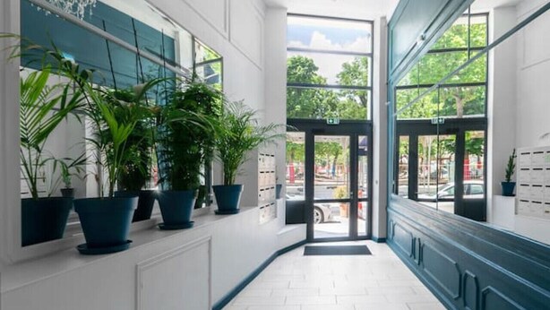 Un pasillo con suelo de baldosas blancas, plantas en macetas, un candelabro y ventanas amplias con vistas al exterior.
