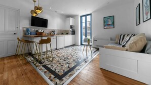 Una cocina moderna con armarios blancos, una zona de comedor con taburetes, una alfombra estampada y un sofá con cojines.