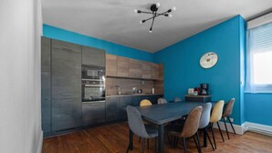 Een moderne keuken met grijze kastjes, een blauwe wand en een eethoek met een tafel en stoelen.