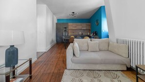 Een modern appartement met een woonkamer, voorzien van een beige bank, een glazen salontafel en een kamerkleed met dessin. De keuken heeft houten kastjes en een blauwe accentmuur.