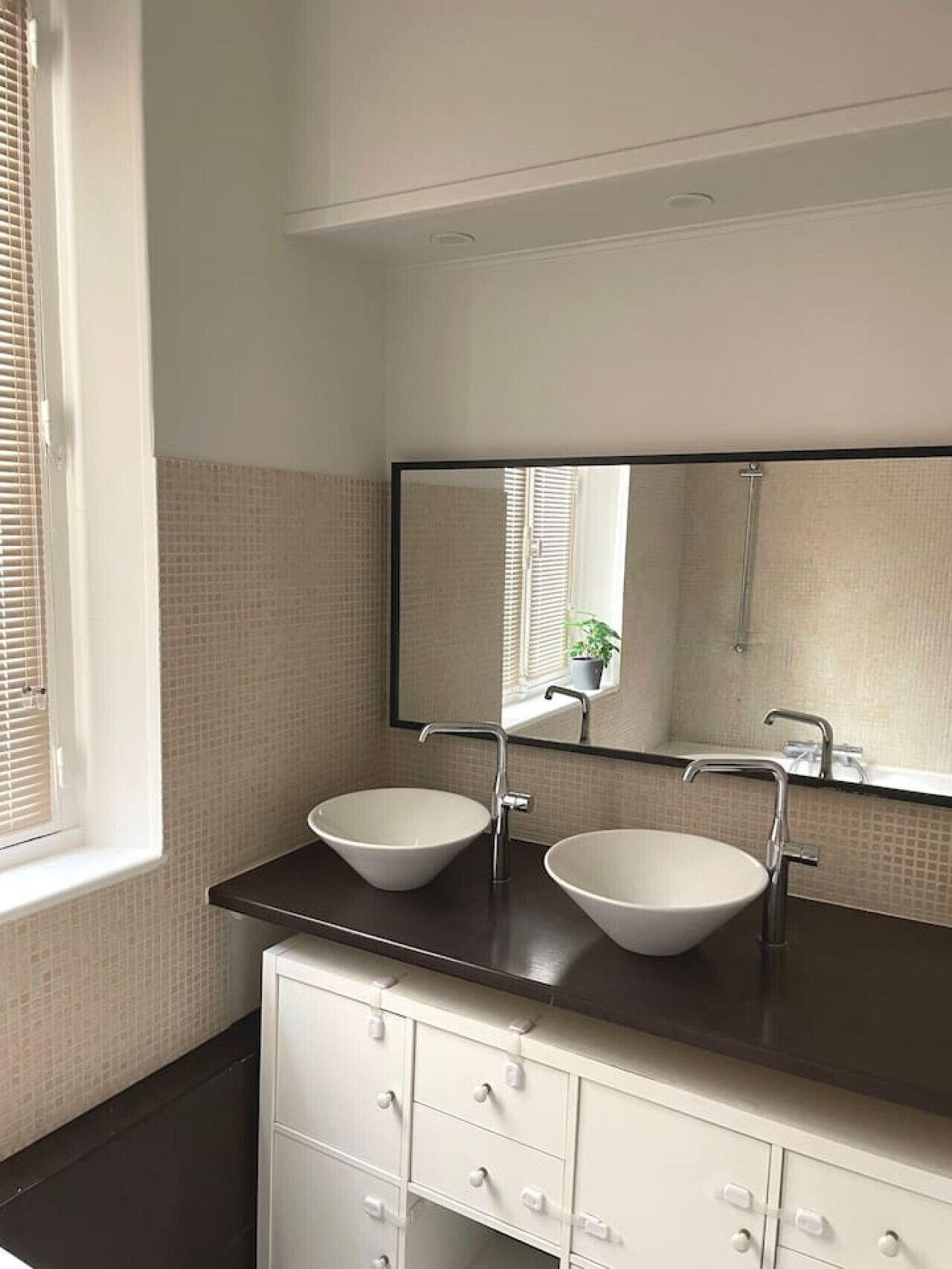 Une salle de bain avec deux vasques blanches, un plan de toilette foncé, un miroir au-dessus des vasques et une fenêtre avec des stores.