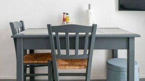 A dining area with a grey table, a wooden chair with a woven seat, a small blue ottoman, and a wall-mounted shelf with a plant and a small item. - Studio design - centre ville - parcs (Vichy)