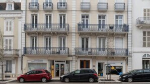 A multi-story building with balconies, a red car parked in front, and a black car parked on the street. - Studio design - centre ville - parcs (Vichy)