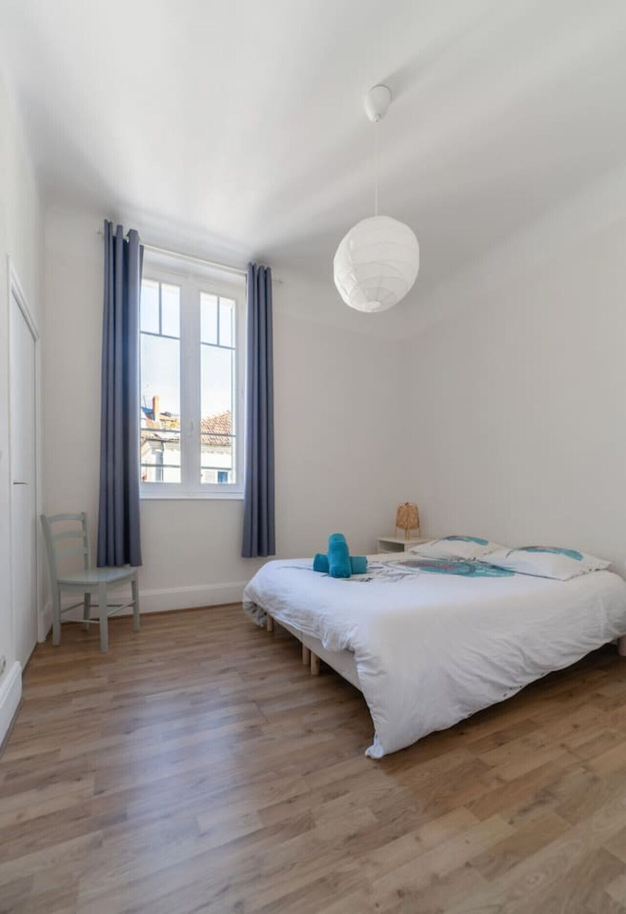 A bedroom with a bed, a chair, a window with curtains, and a hanging light fixture.