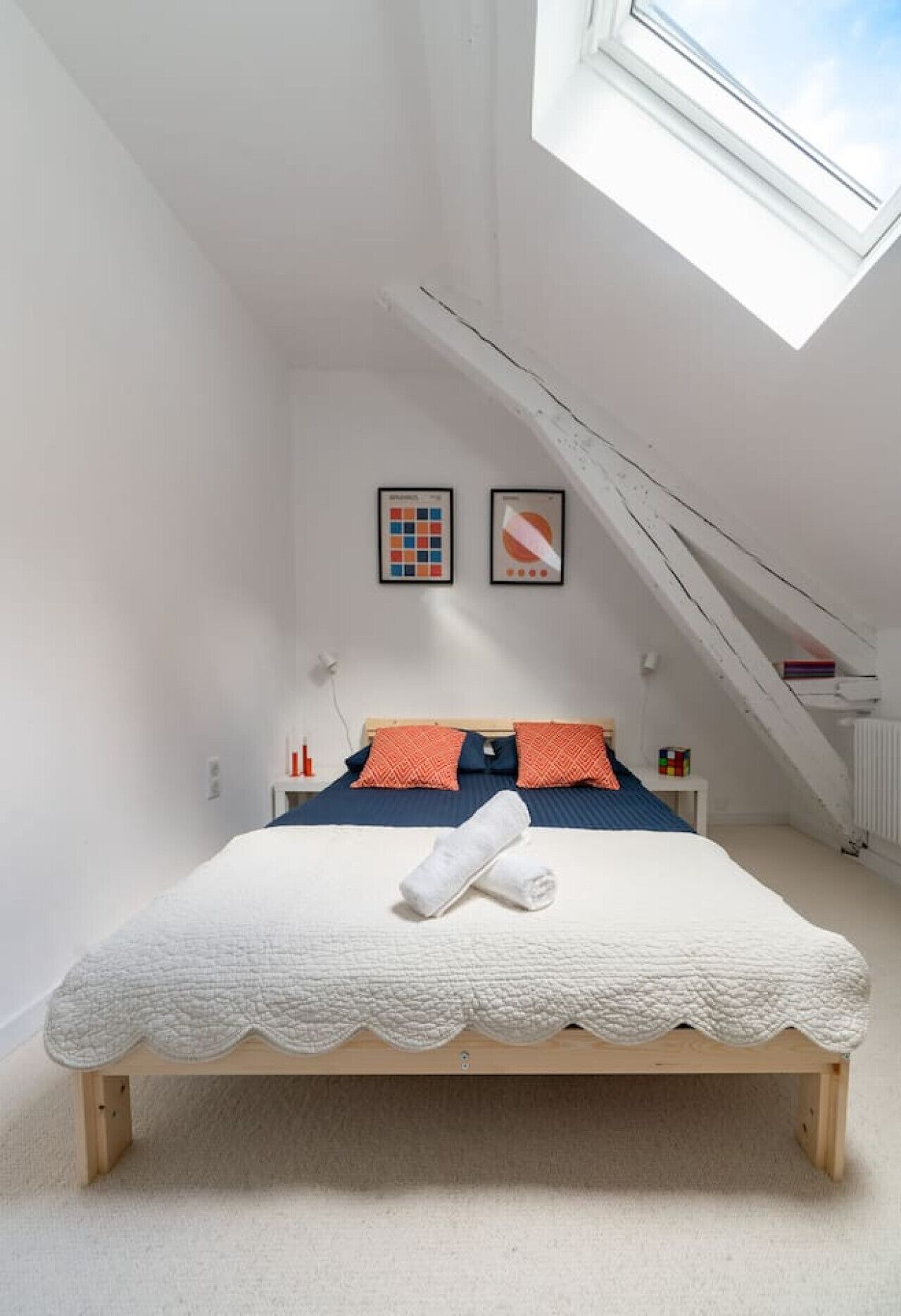 Una camera da letto con soffitto a falde, un letto con copripiumino bianco, due cuscini e un comodino con una bottiglia e una scatola.