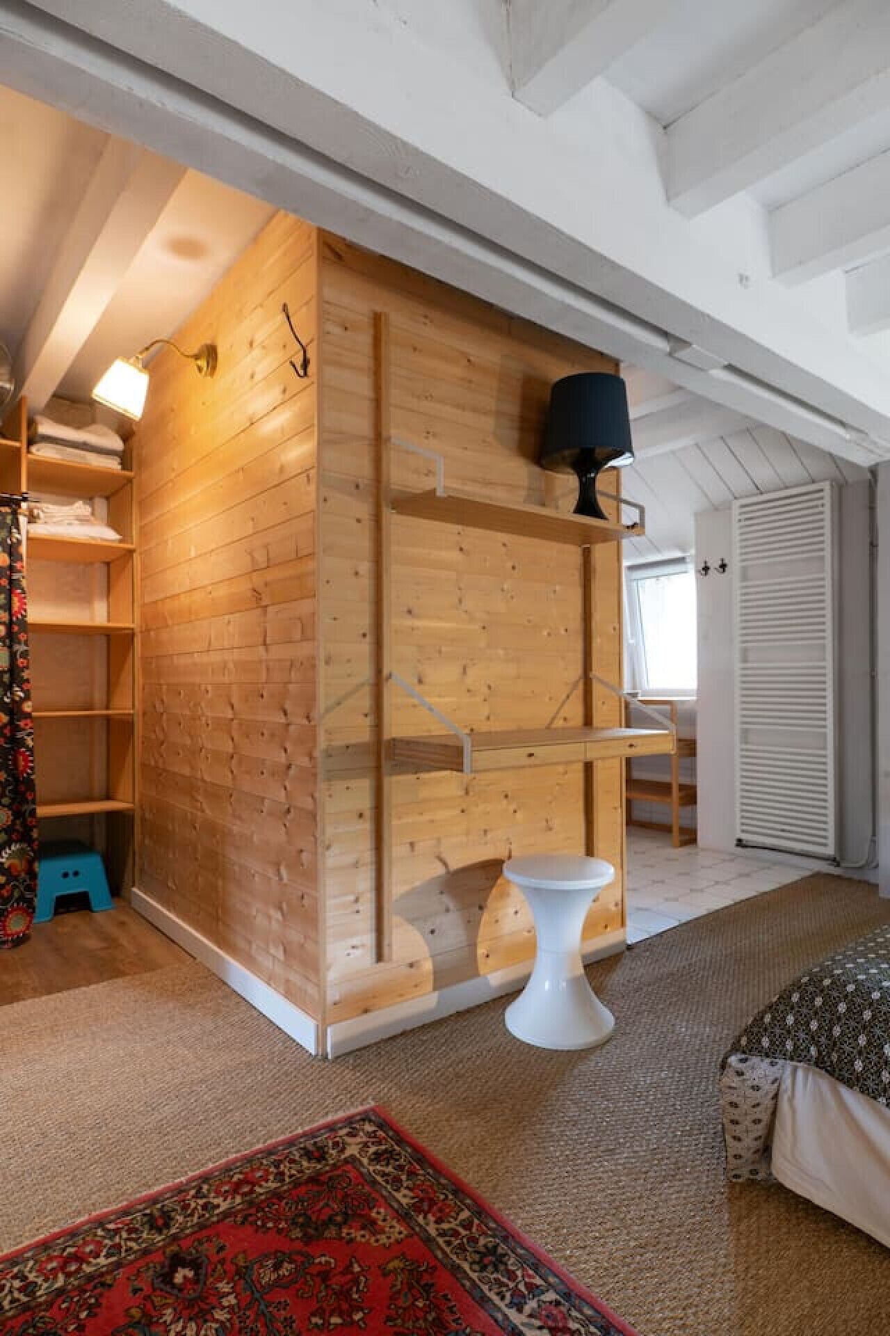 A room with a wooden wall unit, a white pedestal table, a patterned rug, and a bed with a patterned bedspread.