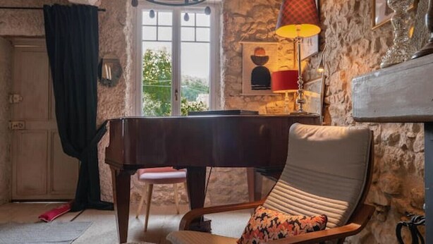 A rustic room with a piano, a cushioned chair, a woven pouf, and a stone wall. - Grande Maison design, terrasse, jardin, parking (Creuzier-le-Neuf)