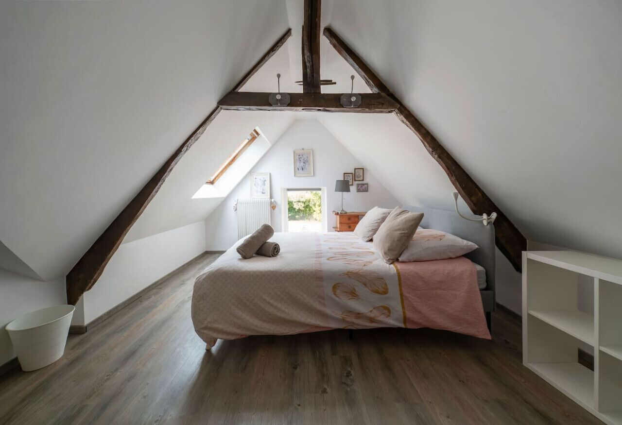 A bedroom with a sloped ceiling, a bed with a pink and white bedspread, a white radiator, and a window with a view of greenery outside.