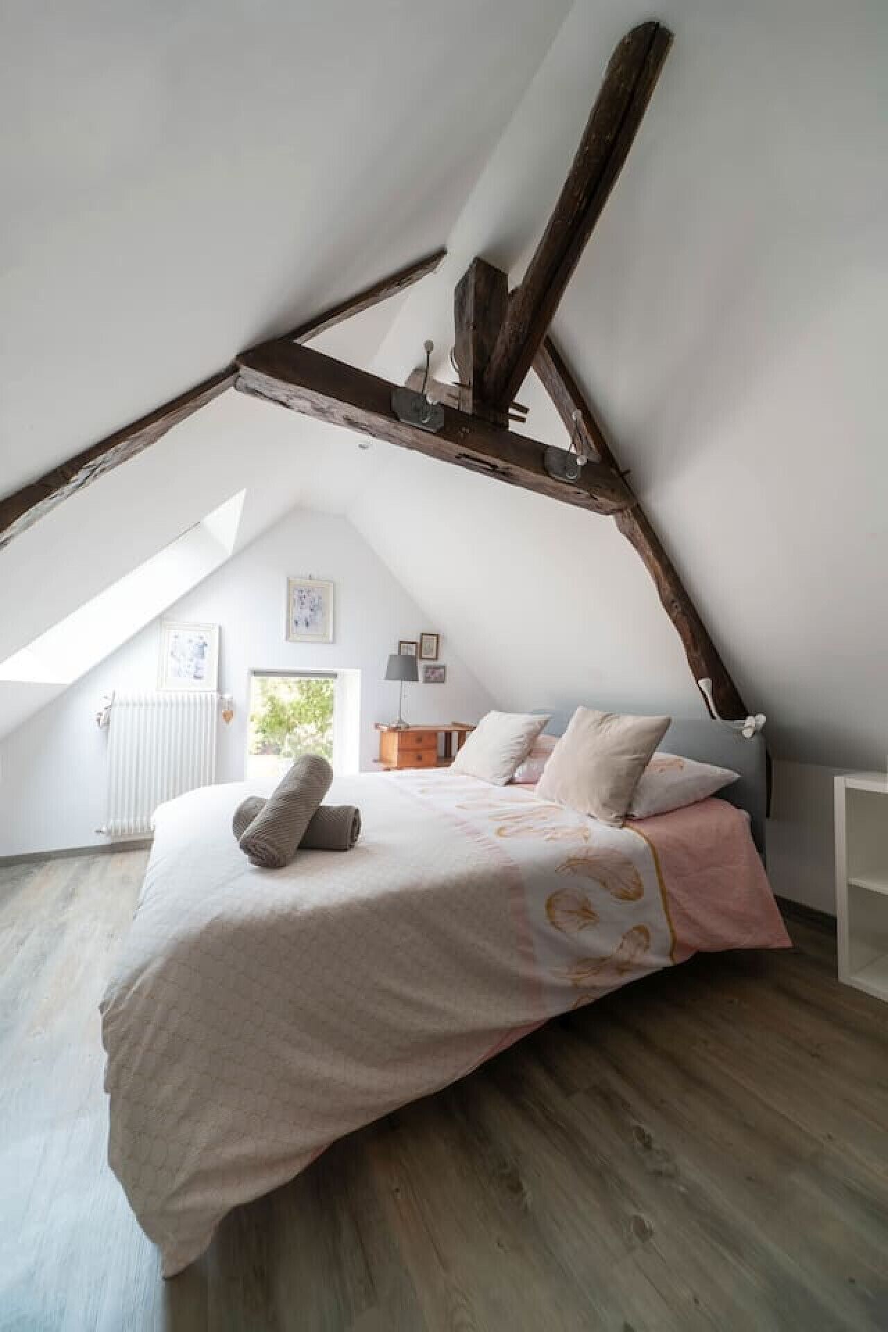 Een slaapkamer met een schuine plafond, houten balken, een bed met een roze en witte spreideken en een raam met uitzicht op groen.