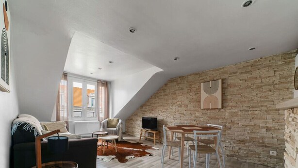 A loft-style room with a sloped ceiling, exposed brick walls, a dining area with a table and chairs, a sofa, a television, and a cowhide rug. - Charmant 2 pièces Centre Ville (Vichy)