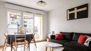 Un soggiorno moderno con angolo da pranzo, un divano nero con cuscini rossi e un'ampia finestra che offre vista sugli edifici.
