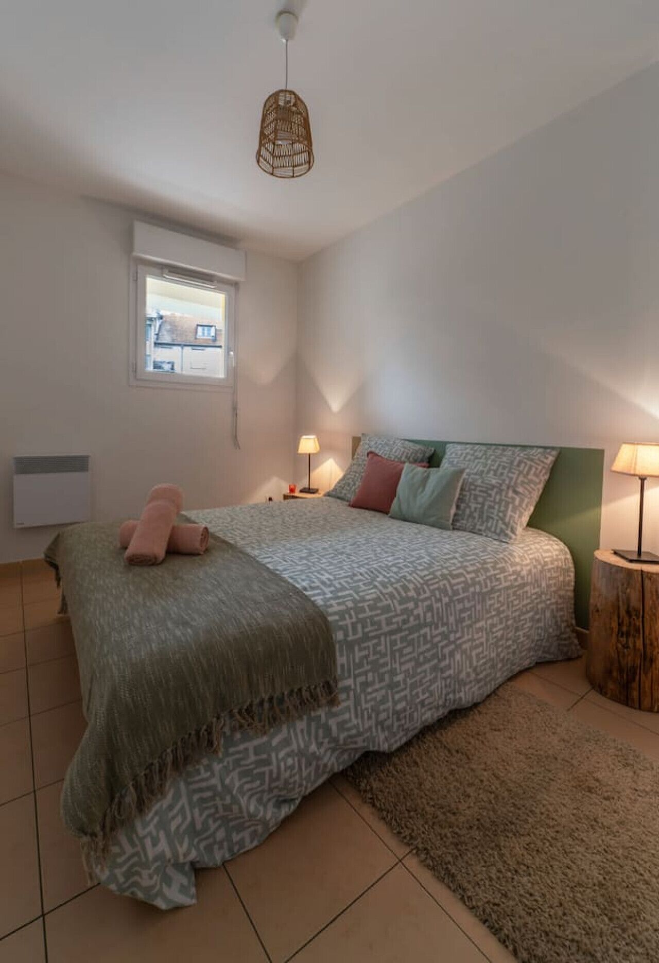 A bedroom with a bed, two bedside tables, a window, a hanging light fixture, and a radiator.