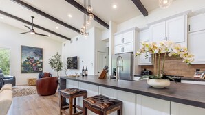 A modern kitchen with white cabinets, a large island with stools, a refrigerator, and a dining area with a sofa and chairs. - Ramsay's Happy Place - River Access & tubes/New Construction (Wimberley)