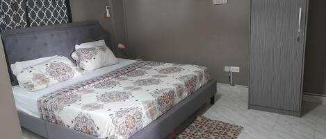 A bedroom with a bed, patterned bedspread, grey headboard, bedside table, lamp, wardrobe, and a patterned rug.