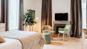 A hotel room with a bed, a TV mounted above a fireplace, a green armchair, a round side table, and a potted plant. - Domitys - Château de la Pilule (Saint-Quentin)