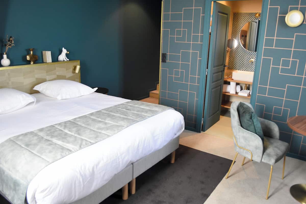 Quarto de hotel com cama, uma cadeira, penteadeira com espelho e uma pequena mesa.