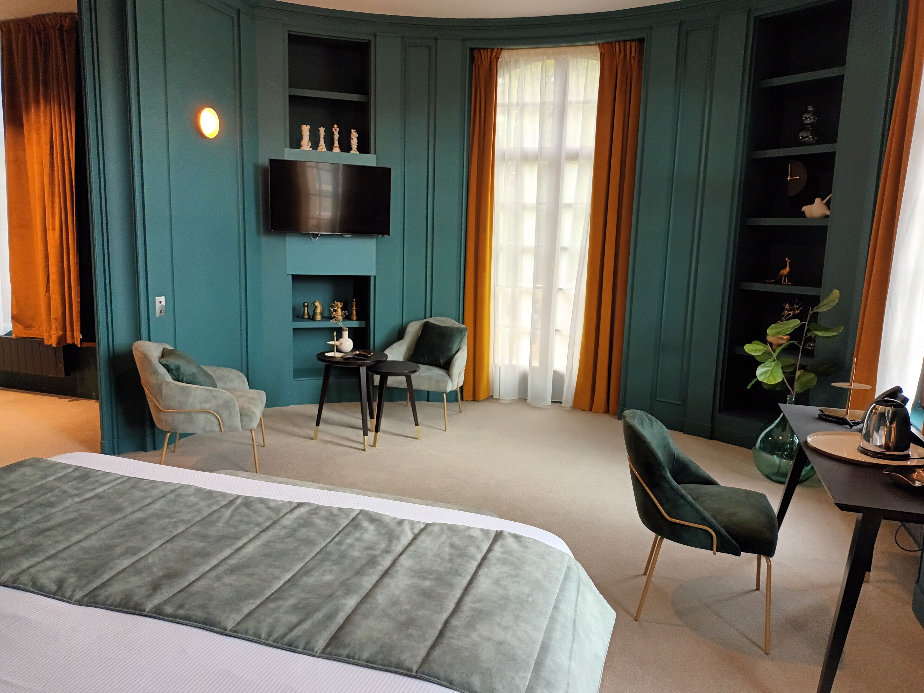Ein modernes Hotelzimmer mit Türkiswänden, einem großen Bett, zwei Sesseln, einem kleinen Tisch und einem an der Wand montierten Fernseher.