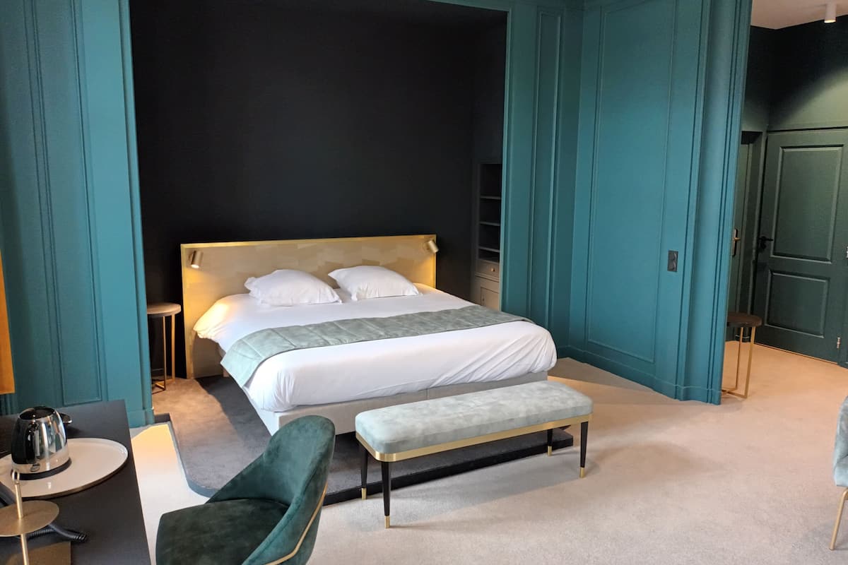 Quarto moderno com uma cama grande, um banquinho, uma cadeira e uma pequena mesa com uma chaleira.