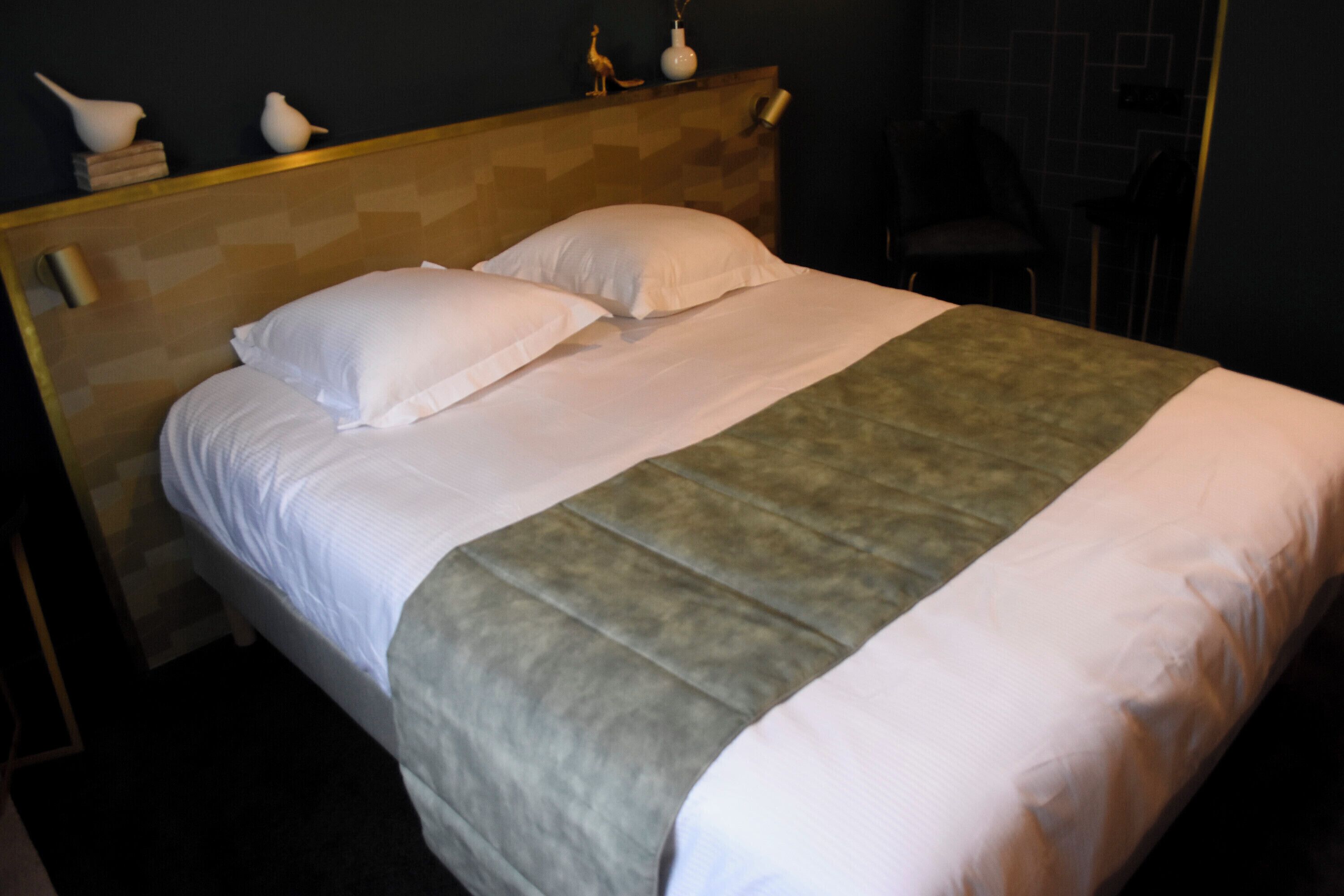A neatly made bed with white linens and a grey accent throw, a wooden headboard, and a small shelf with decorative items.