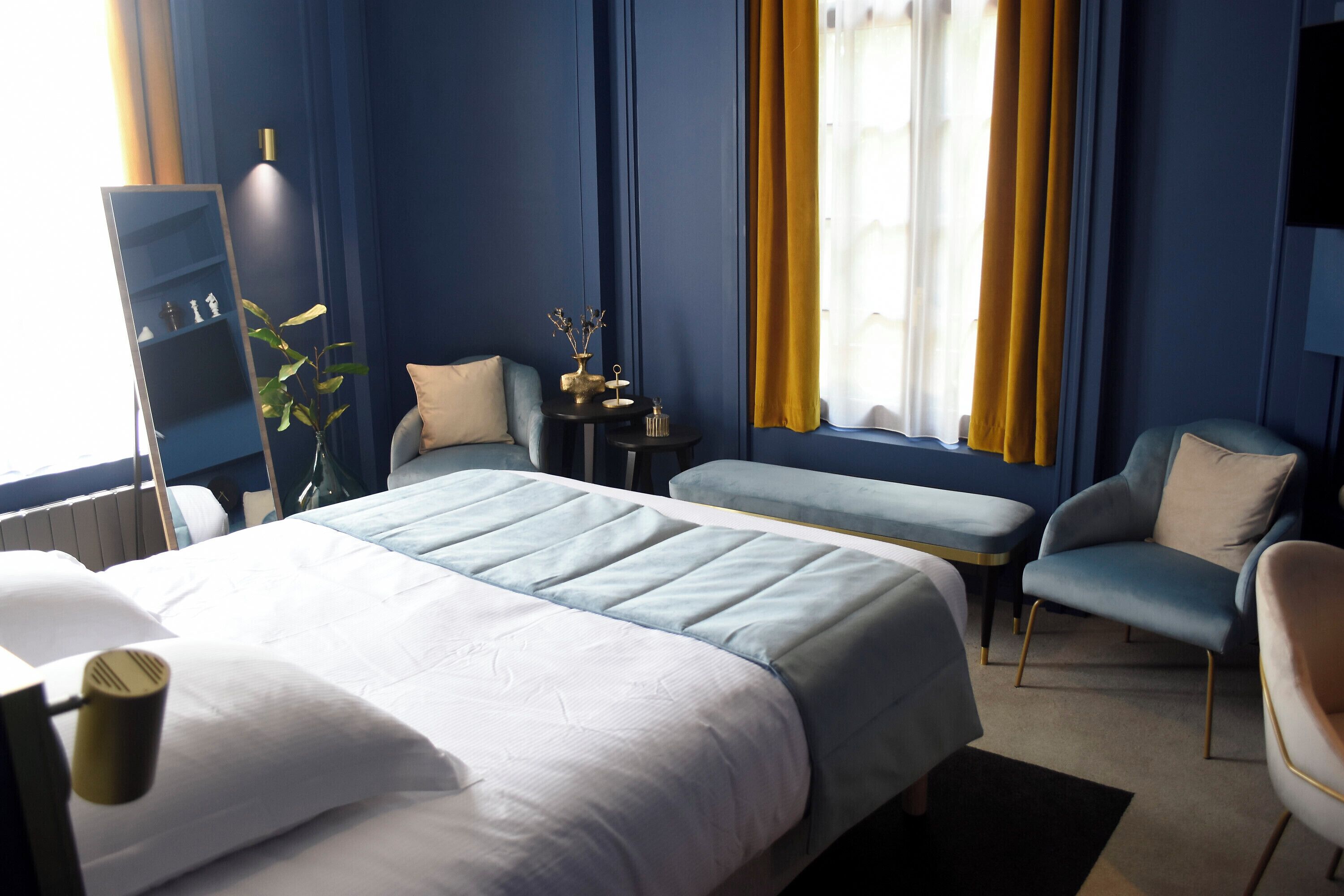 Une chambre d’hôtel avec un lit, deux fauteuils, une petite table, un miroir et une plante.