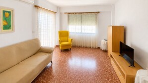 Een kamer met een bank, een gele fauteuil, een televisie op een houten dressoir en een raam met gordijnen.