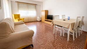 Een kamer met een bank, een eettafel met stoelen, een televisie op een dressoir en een raam met gordijnen.