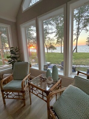 Una habitación con ventanas amplias, mobiliario de mimbre y vistas a un atardecer sobre el agua.