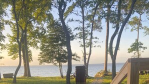 A bottle of Meomi Pinot Noir wine on a table with two glasses, set outdoors with a view of trees and a body of water in the background.