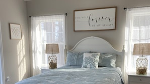 A bedroom with a bed, two bedside tables, a wall-mounted sign, and a window with sheer curtains.