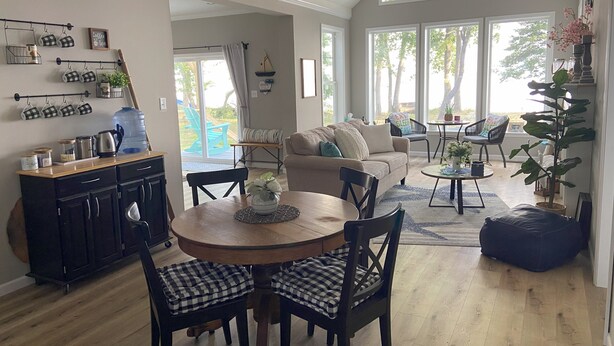 A spacious living area with a large window, a dining table with chairs, a sofa, and a kitchen area with a water dispenser and mugs.