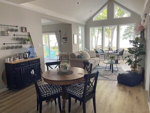 Un amplio salón con un gran ventanal, una mesa de comedor con sillas, un sofá y una zona de cocina con dispensador de agua y tazas.