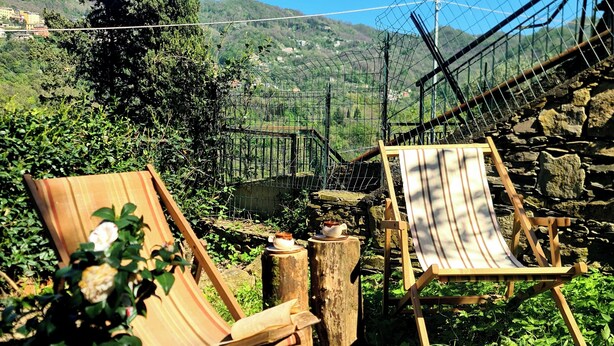 Un espace extérieur aménagé dans un style rustique, avec des chaises et une table en bois, entouré de verdure et bordé d’un mur en pierre.