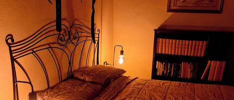 Een kamer met weinig licht, met een bed, nachtkastje, lamp en een boekenplank.