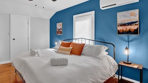 A bedroom with a bed, a ceiling fan, an air conditioner, and two wall-mounted artworks.