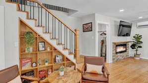 A living room with a wooden staircase, a fireplace, a television mounted on the wall, and a bookshelf filled with books and decorative items.