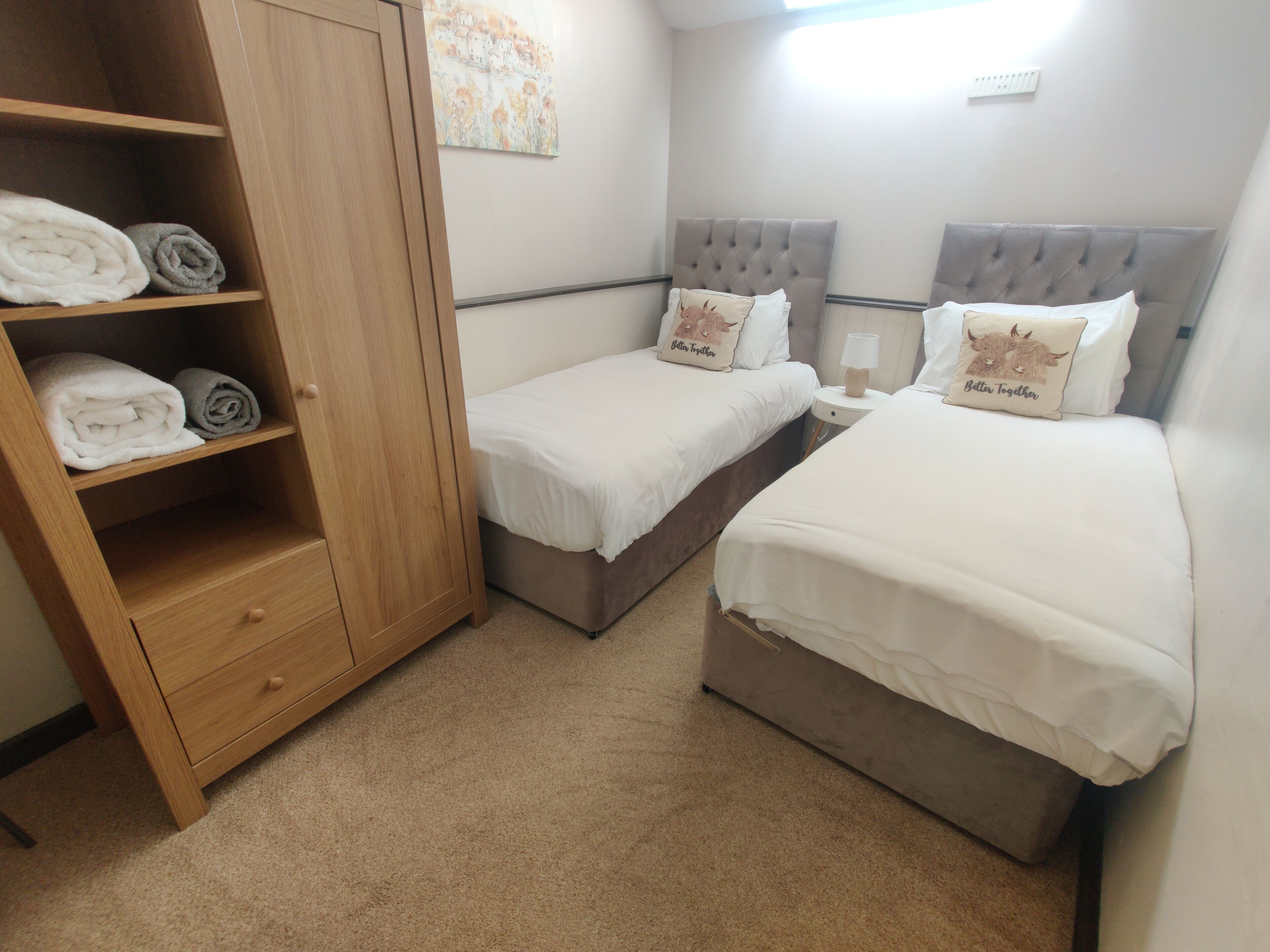 A bedroom with two single beds, a wooden wardrobe, and a small bedside table with a lamp.
