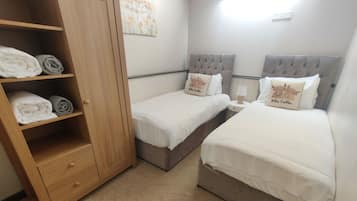 A bedroom with two single beds, a wooden wardrobe, and a small bedside table with a lamp.