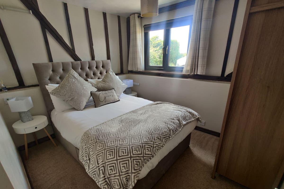 Quarto com cabeceira estofada, colcha estampada, mesa de cabeceira com luminária e janela com cortinas.