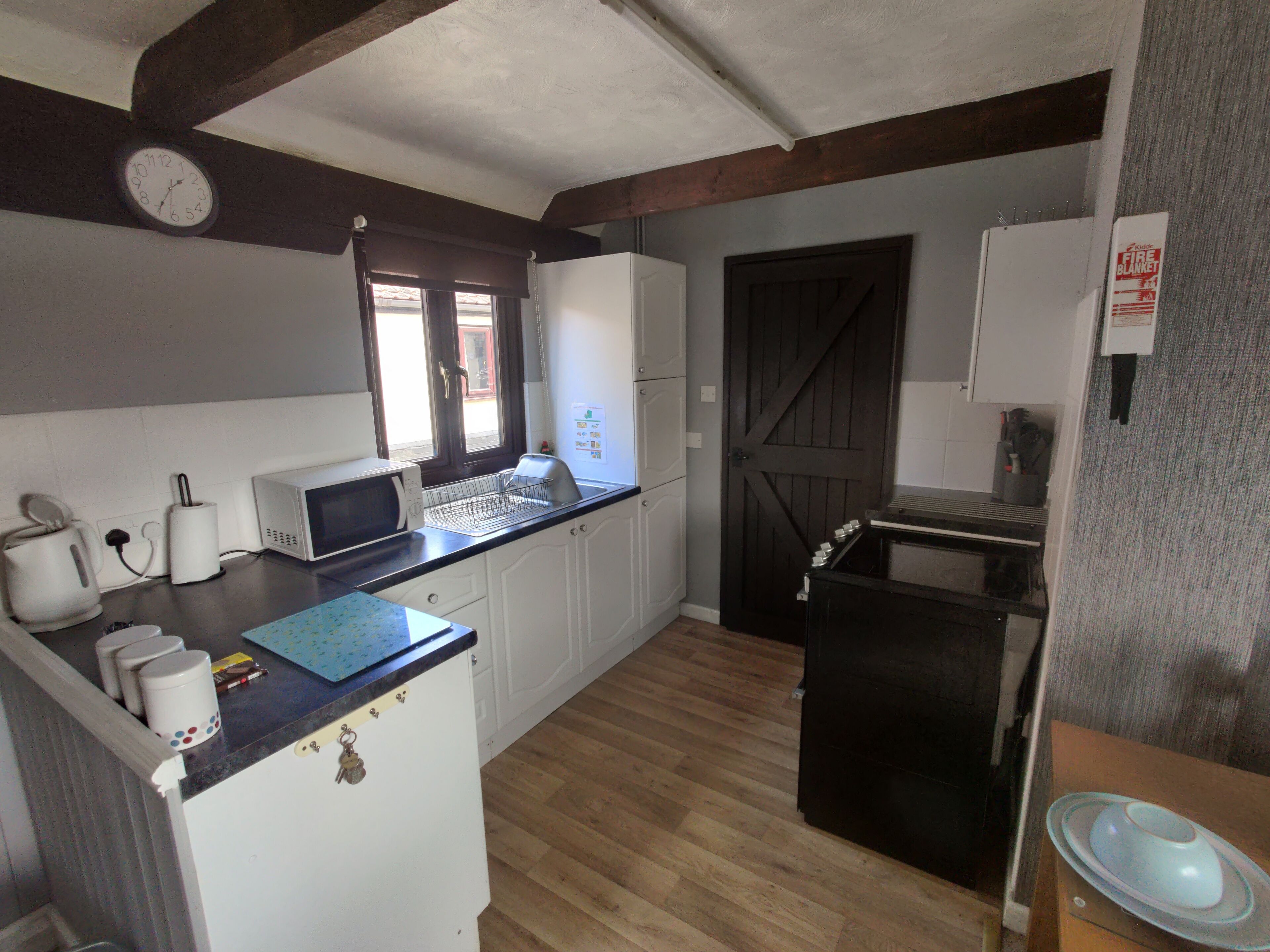 Eine kleine Küche mit Mikrowelle, Toaster und Spüle. Ein Holztür und ein Fenster mit Jalousien sind vorhanden.